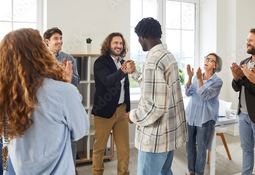 Diverse business team celebrating success and having fun in office. Two young men exchange handshakes while group of happy joyful mixed race colleagues clap hands and cheer. Teamwork success concept