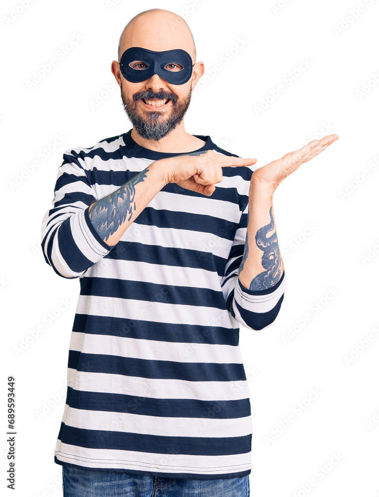 Young handsome man wearing burglar mask amazed and smiling to the camera while presenting with hand and pointing with finger. Young handsome man wearing burglar mask amazed and smiling to the camera while presenting with hand and pointing with finger.