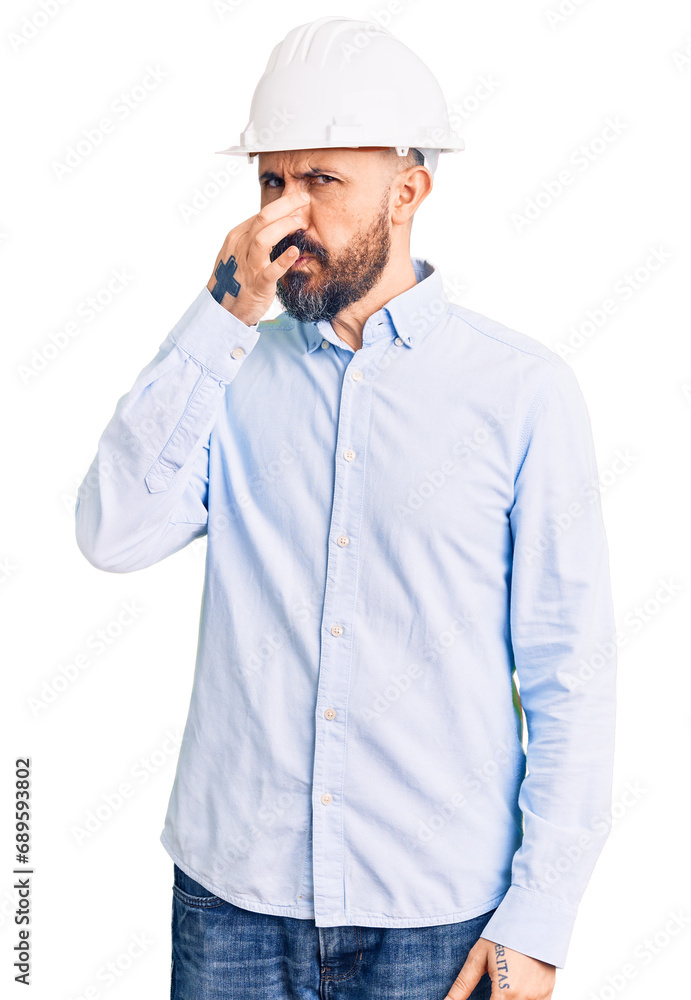 Young handsome man wearing architect hardhat smelling something stinky and disgusting, intolerable smell, holding breath with fingers on nose. bad smell Young handsome man wearing architect hardhat smelling something stinky and disgusting, intolerable smell, holding breath with fingers on nose. bad smell