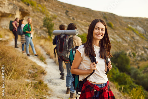 Group of active friends hiking or trekking on mountain trail on warm autumn weekend. Happy young woman hiker standing on hill pathway with blurry landscape in background, looking at camera and smiling