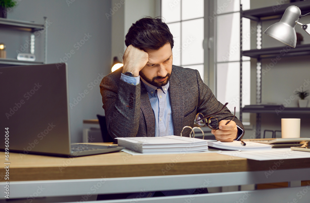 Auditor or accountant gets very tired while doing paperwork in the office. Young man in a suit sitting at his working desk with a thick accounting book, leaning his head on his hand and thinking Auditor or accountant gets very tired while doing paperwork in the office. Young man in a suit sitting at his working desk with a thick accounting book, leaning his head on his hand and thinking