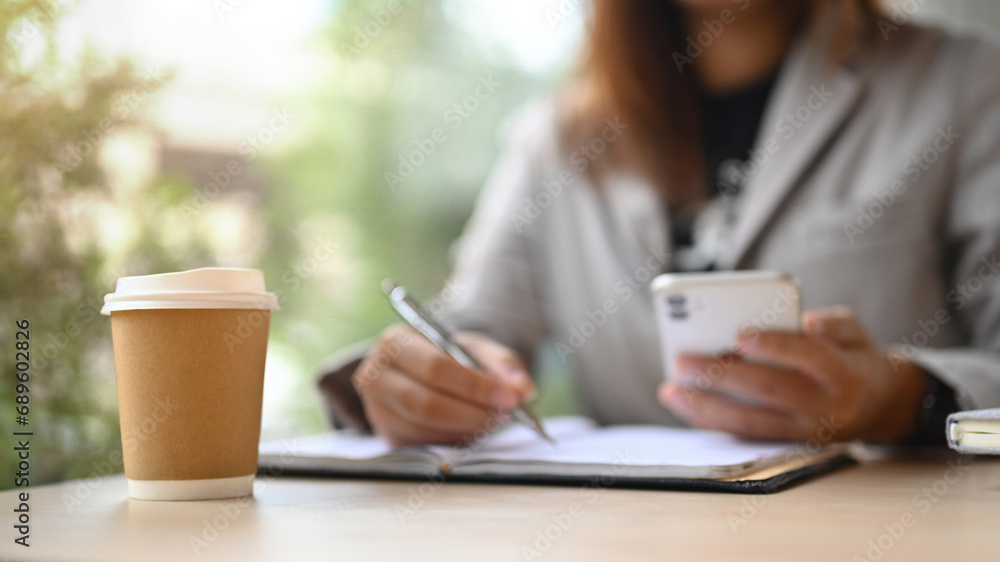 A paper cup of coffee on wooden office desk with businesswoman sitting on background A paper cup of coffee on wooden office desk with businesswoman sitting on background