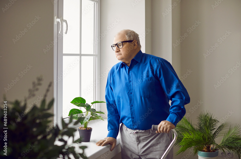 Portrait of pensive senior man looking at window. Lonely thoughtful elderly man with walking cane standing near window at home. Aging, loneliness, disability concept Portrait of pensive senior man looking at window. Lonely thoughtful elderly man with walking cane standing near window at home. Aging, loneliness, disability concept