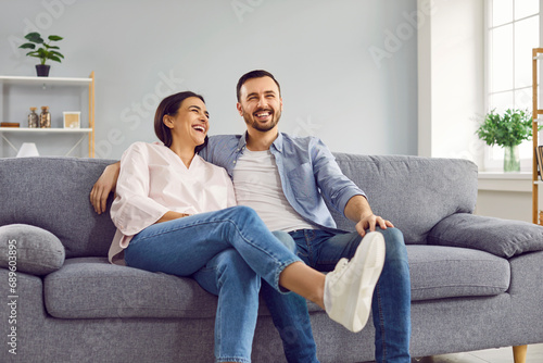 Young family couple having fun at home. Happy husband and wife sitting on a comfortable grey couch and laughing. Cheerful husband and wife sitting on the sofa and laughing at a funny joke