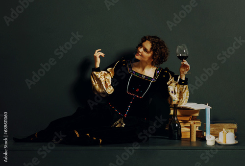 Woman dressed in a medieval dress sitting near ancient books and candles with bottle of red wine and holding a glass of wine. Old oil painting stylization