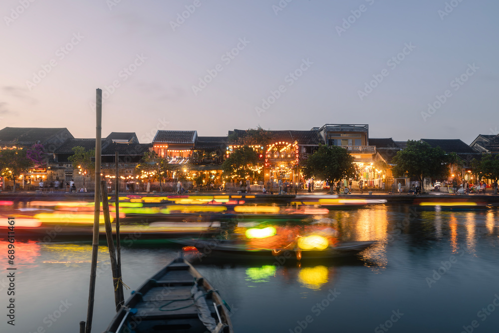 Traditional boats with lanterns in long exposure against illuminated busy waterfrost of ancient city. Old town in Hoi An, popular tourist destination in Vietnam..