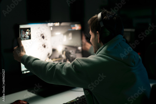 Back view of male cybersport gamer in headset touching computer monitor while celebrating successful game in dark room
