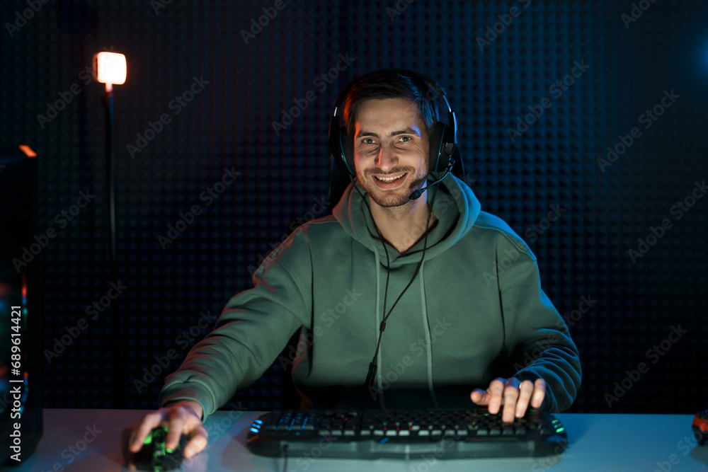 Smiling young male cybersport gamer in hoodie and headset using keyboard and mouse while playing video game in dark room looking at camera Smiling young male cybersport gamer in hoodie and headset using keyboard and mouse while playing video game in dark room looking at camera