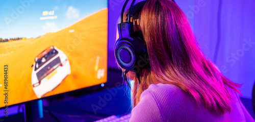 Happy Gamer endeavor plays online video games tournament with computer with neon lights, Young player woman wearing gaming headphones intend to do playing car games online at home, Back view
