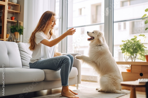 Young woman playing with fluffy dog in bright living room. Obedience training and cuddling with pets at home.