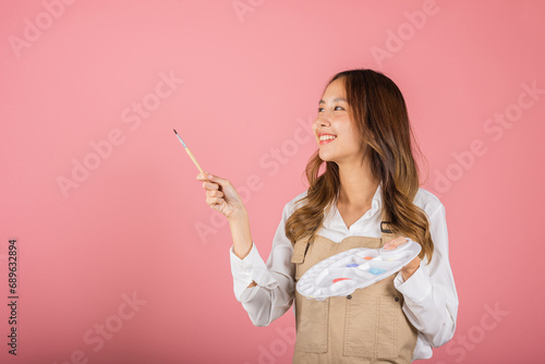 Asian beautiful young woman artist holding brush and paint palette, Happy female painting using paintbrush and palette with colors, studio shot isolated on pink background, Paintings and art equipment photo