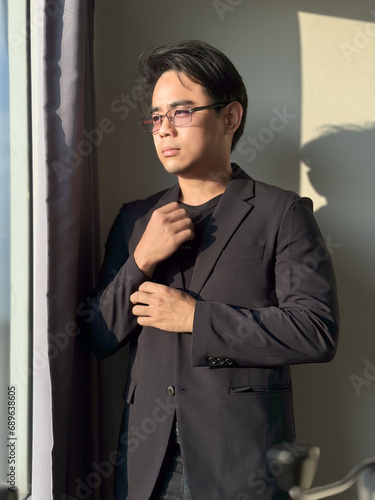 Vertical portrait of an Asian man with short, cool black hair, glasses, wearing a black business suit, buttoning the sleeves of his shirt. The background is the shadow of a window.