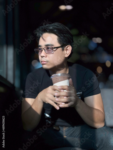 vertical portrait of an Asian man with short, cool black hair, wearing glasses, wearing a black shirt, is drinking coffee with a cafe in the background.