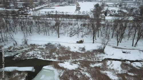A drone monitors a tractor driving along a snowy road along a river in an industrial area in winter.