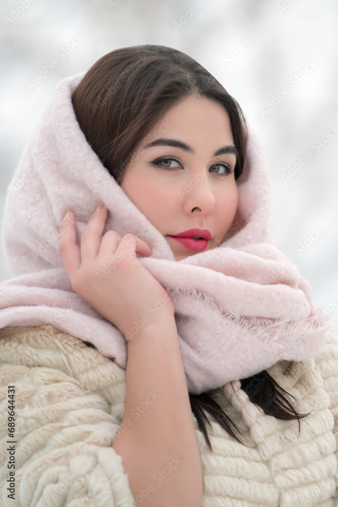 Obraz premium Winter portrait of brunette with red plump lips, dressed in white fur coat and light pink scarf on head. Soft lighting on elegant young adult female model on evening. Depth of field, selective focus