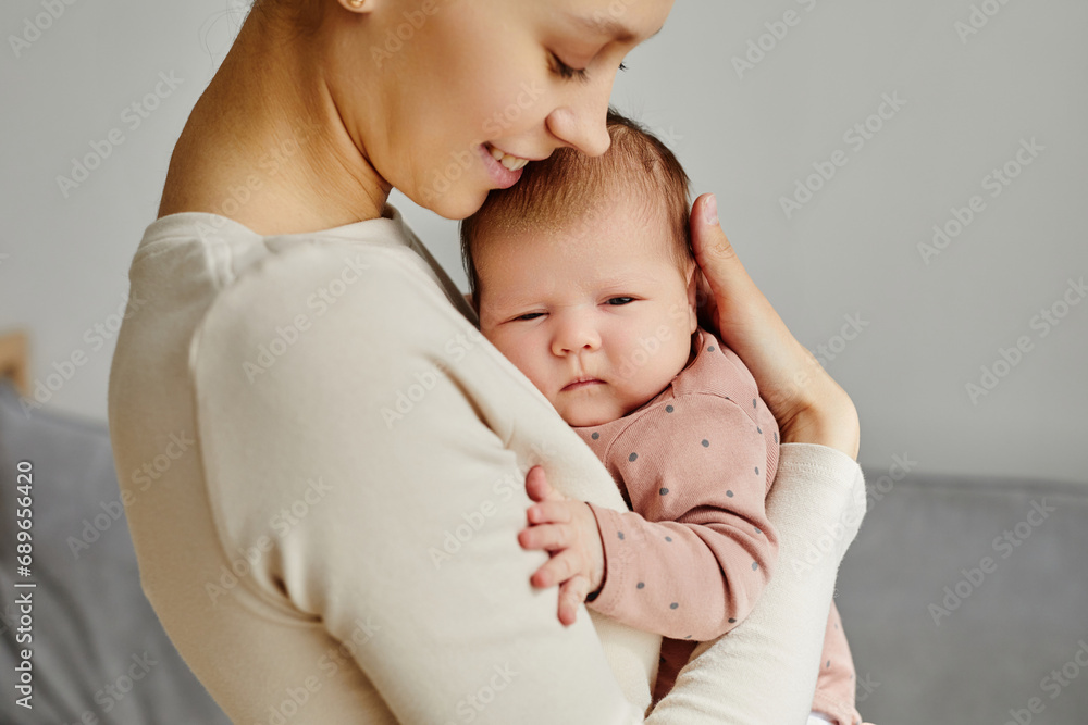 Side view at young smiling Caucasian woman hugging her sleepy newborn looking at camera, copy space Side view at young smiling Caucasian woman hugging her sleepy newborn looking at camera, copy space
