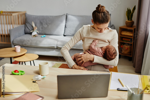 Medium shot of young woman breastfeeding infant while sitting at home office