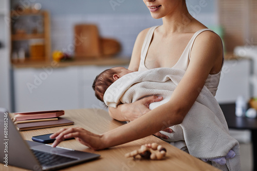 Cropped shot of unrecognizable woman working on computer and hugging her sleeping baby in soft blanket, copy space