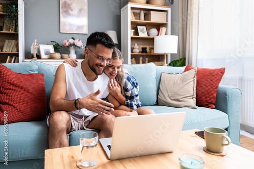 Happy young couple at home man surprise woman to choose travel destination, searching the internet on laptop computer. Online booking. Anniversary vacation and celebration of togetherness and love photo