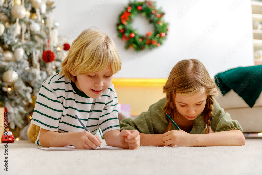 boy and girl write together letters with wish list to santa claus using color pencils laying on floor near christmas tree. boy and girl write together letters with wish list to santa claus using color pencils laying on floor near christmas tree.