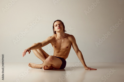Handsome young guy with sportive, muscular body sitting on floor, posing shirtless in underwear against grey studio background. Concept of men's beauty, health, body care, sportive lifestyle