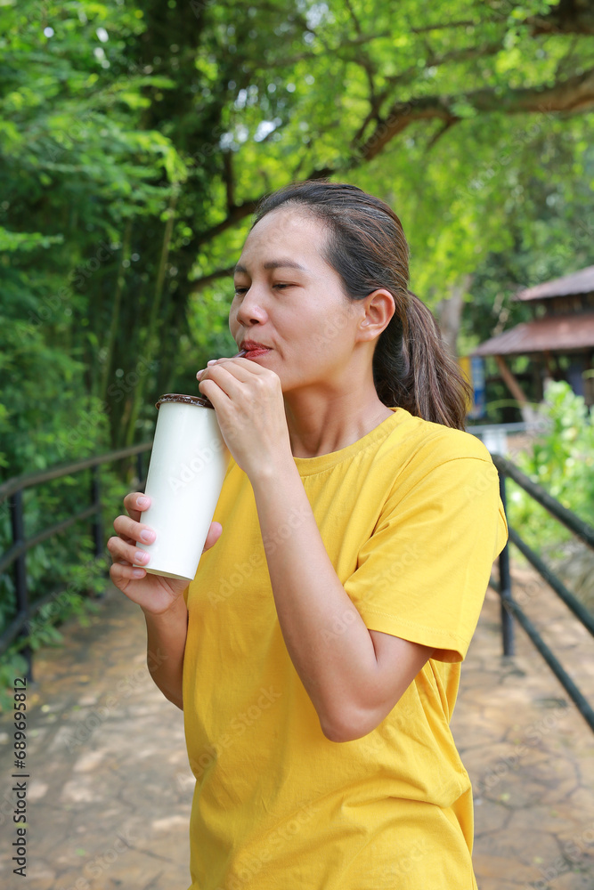 Portrait of Asian woman holding paper cup and drinking cold water by straw while standing in public garden. Portrait of Asian woman holding paper cup and drinking cold water by straw while standing in public garden.