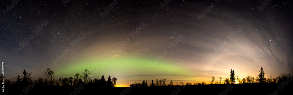 Panoramic view of the night sky with many stars, the Aurora along the horizon and the moon rising on the right side.  Mixed trees are silhouetted along the horizon.
