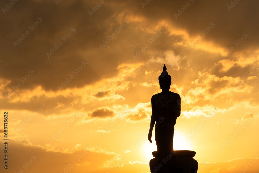 Stand big Buddha Statue in silhouette sun set Light background in park of Thailand temple.Yellow orange light silhouette dark shadow of image Buddha statue stand Buddha tall walk in sun light cloud.