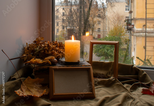 Cuadro en lienzo Empty wooden frames for photos or paintings, a burning candle on a stack of old books and dry autumn leaves on the windowsill against the backdrop of a window with an urban landscape