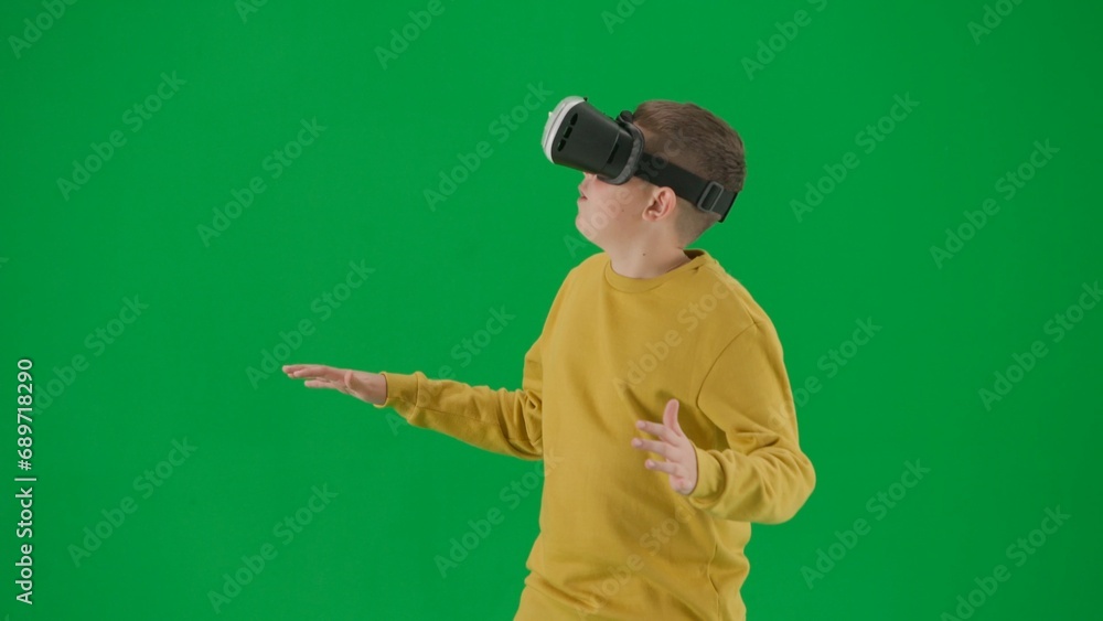 Portrait of kid boy on chroma key green screen. Schoolboy in jeans wears virtual reality glasses amazed face expression. Middle body front shot.