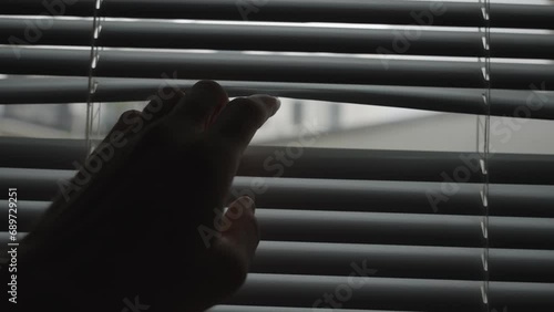 Hand gets close to shut window blinds and crack opens one to peek outside room. Dark office room or house with hand reaching towards window to spy and watch and look out from inside apartment