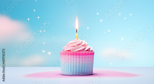 A pink and white cupcake with a lit candle on top. The cupcake is in a pink paper liner with a white swirl of frosting on top. The background is a bright blue with small pink and blue sprinkles.