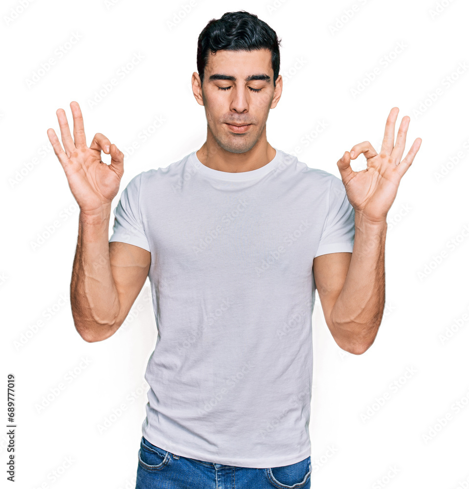 Handsome hispanic man wearing casual white t shirt relax and smiling with eyes closed doing meditation gesture with fingers. yoga concept. Handsome hispanic man wearing casual white t shirt relax and smiling with eyes closed doing meditation gesture with fingers. yoga concept.