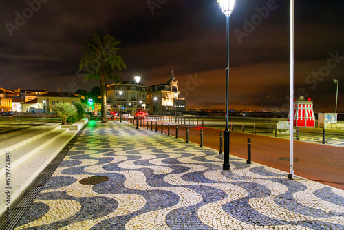 A traditional calçada pavement pattern at night along the seaside promenade at Praia da Rainha, with an illuminated Palacete Seixas Palace, in Cascais Portugal. 