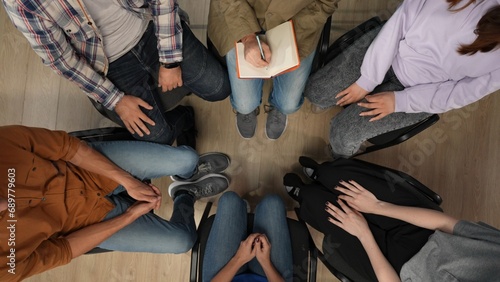 In the shot above a close up of a group of people sitting in a circle with their arms folded. One of them in the center is writing something in a notebook. Imitating a mentor, a doctor in session