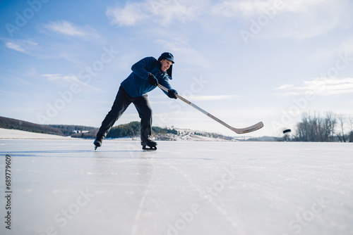 An elderly man practices stricking the puck with hockey stick on a frozen lake in winter.