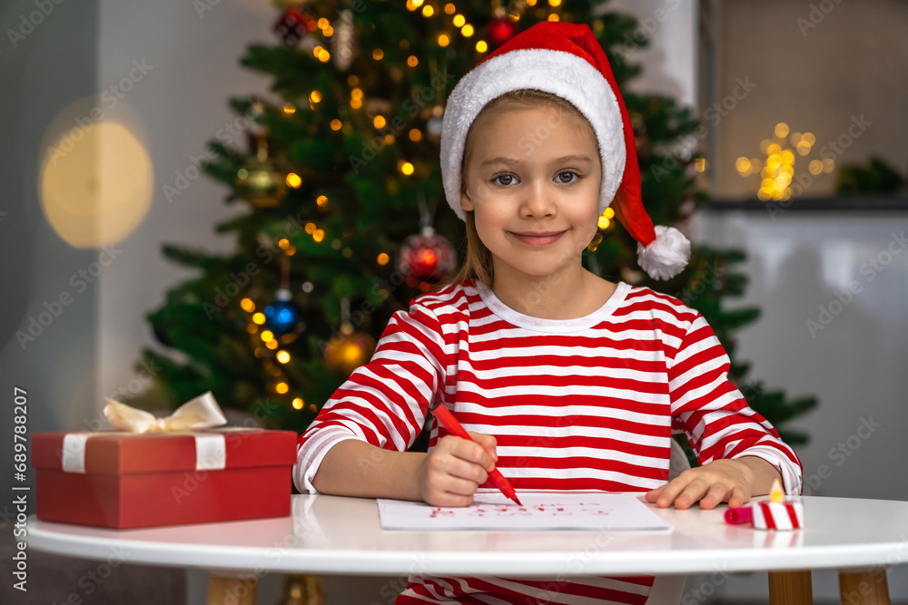 Portrait of happy little girl looks at camera and writes letter to Santa Claus on background of christmas tree. New Year Portrait of happy little girl looks at camera and writes letter to Santa Claus on background of christmas tree. New Year