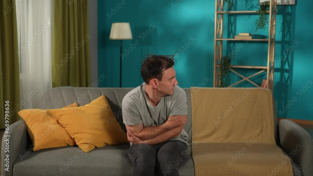 A mentally unstable man is sitting on a sofa in the living room, looking over one shoulder or the other, talking to himself. The unhappy man is clutching his head. Split personality. A mentally unstable man is sitting on a sofa in the living room, looking over one shoulder or the other, talking to himself. The unhappy man is clutching his head. Split personality.