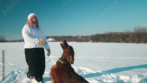 Happy beautiful young woman blowing snowflakes from her hands to her dog irish setter enjoying winter snow outdoors activity in winter day holiday. People and animals together having fun and smiling photo