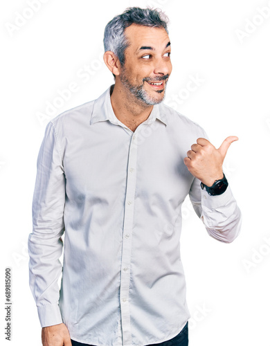 Middle age hispanic with grey hair wearing casual white shirt smiling with happy face looking and pointing to the side with thumb up.