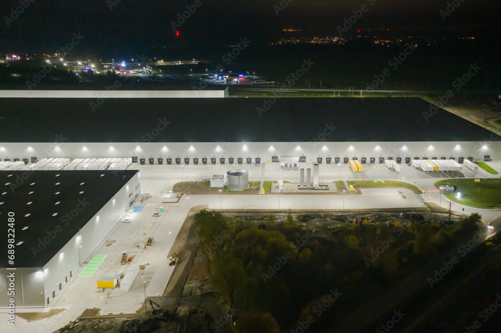 Centrum logistyczne w nocnych ciemnościach. Teren wokół jak i budynki oświetlone są sztucznym światłem elektrycznym. Zdjęcie zrobione przy użyciu drona.