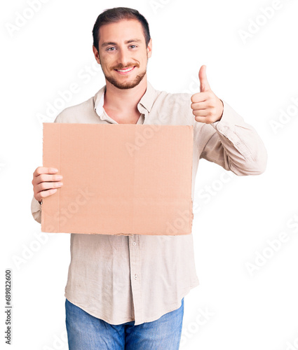 Young handsome caucasian man holding empty cardboard banner smiling happy and positive, thumb up doing excellent and approval sign