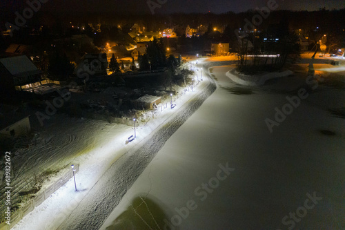 Staw nocą, w zimowej scenerii. Powierzchnnia stawu skuta lodem, brzeg pokryty warstwą białego śniegu. Wokół stawu znajduje się ścieżka przy której stoją ławki i latarnie. Zdjęcie z drona.