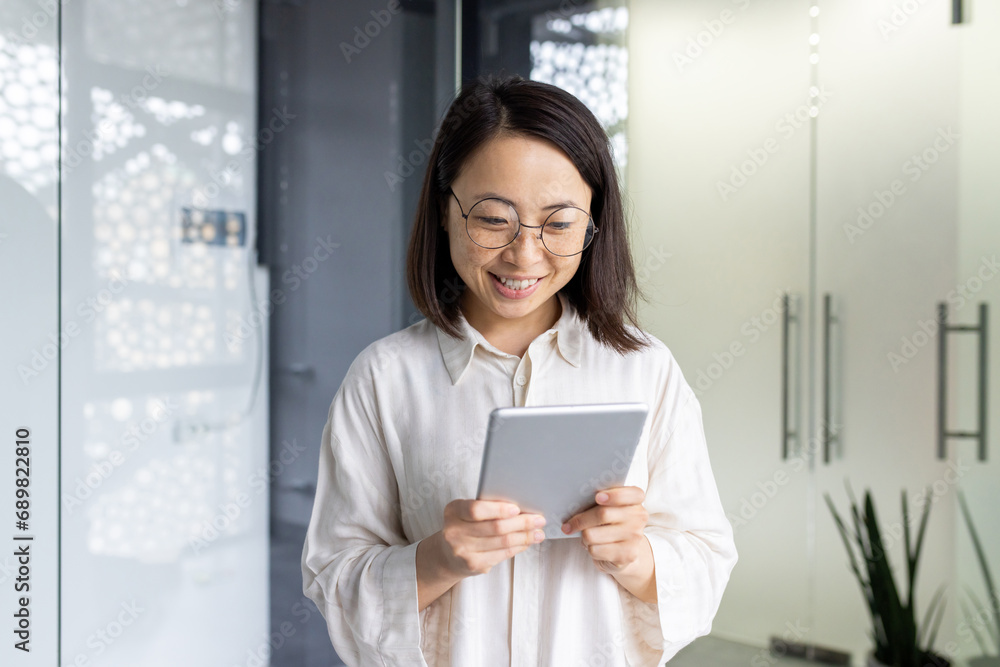Young beautiful Asian woman with tablet computer standing near window, smiling businesswoman using online application, programmer testing new software.