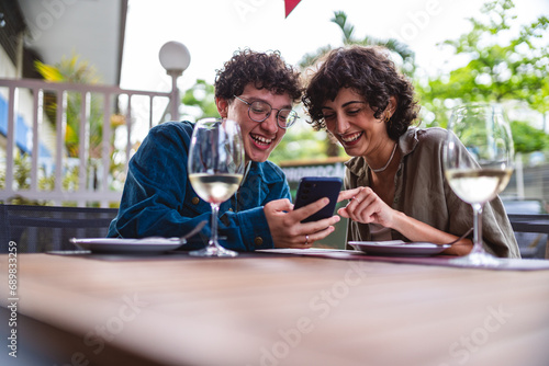 Jovem casal sentado a mesa de um restaurante ao ar livre conversando, usando smartphone. photo