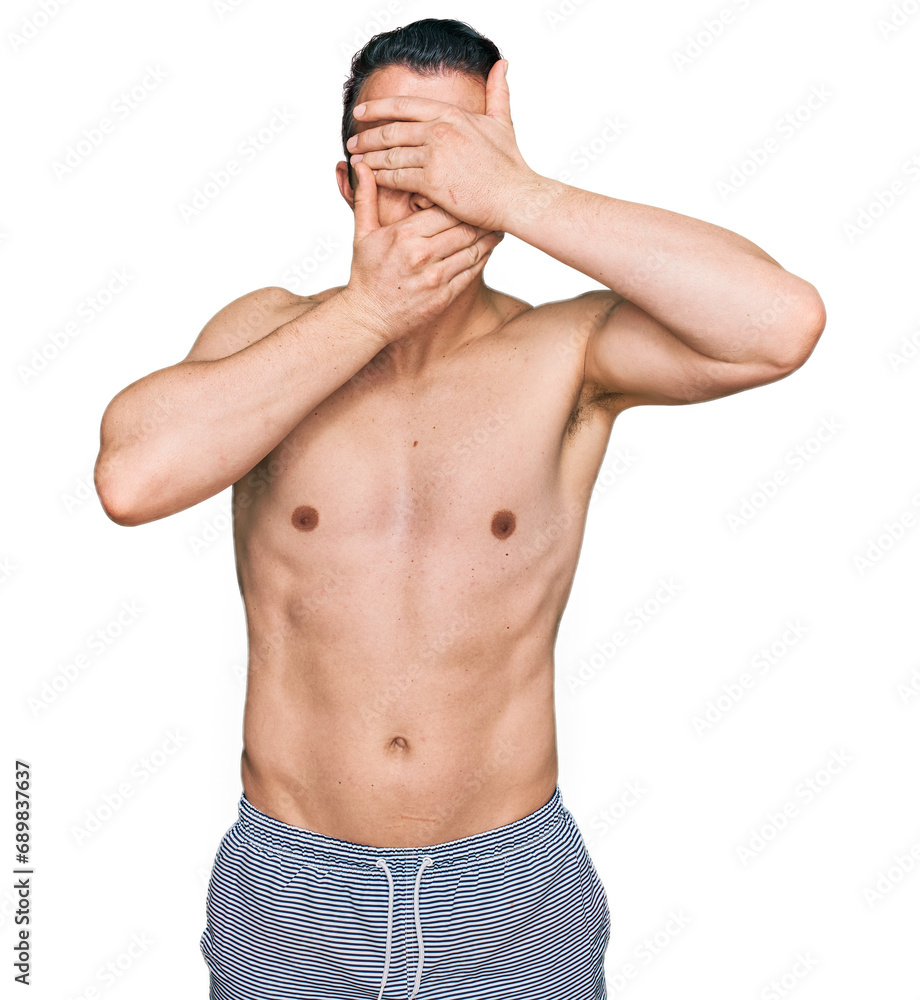 Fototapeta premium Handsome young man wearing swimwear shirtless covering eyes and mouth with hands, surprised and shocked. hiding emotion