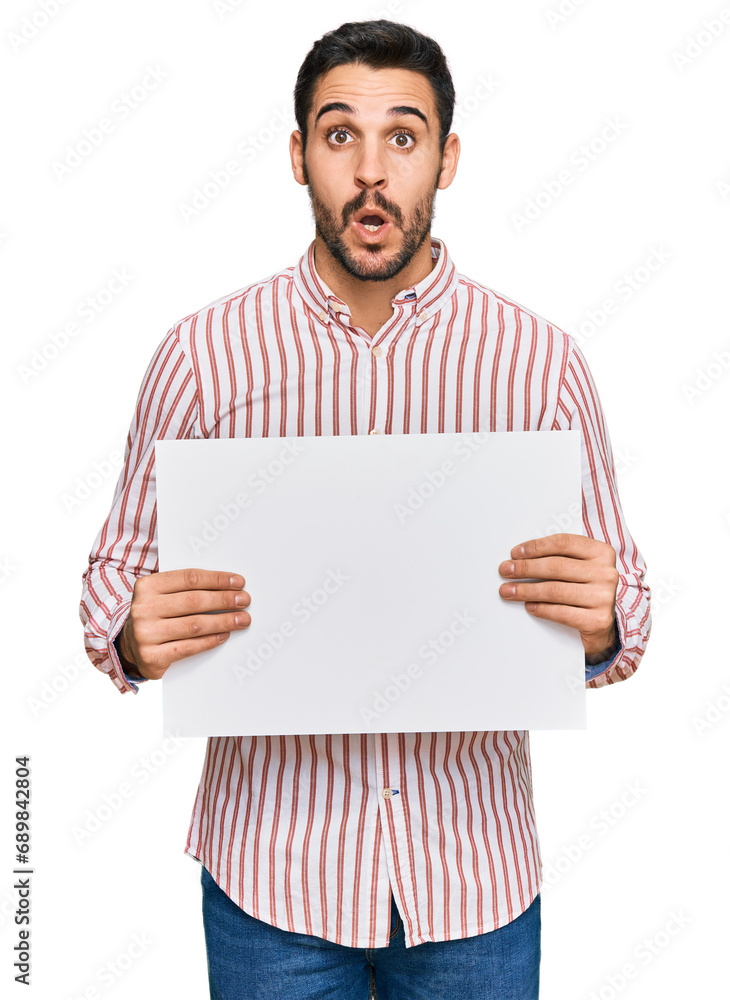 Young hispanic man holding blank empty banner scared and amazed with open mouth for surprise, disbelief face Young hispanic man holding blank empty banner scared and amazed with open mouth for surprise, disbelief face
