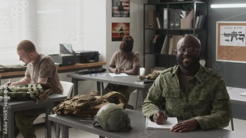 Wide slowmo portrait of smiling male African American cadet in glasses and camouflage uniform studying during class and then looking at camera in military academy