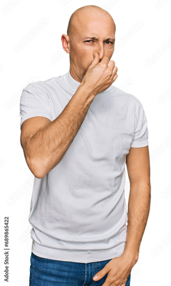 Bald man with beard wearing casual white t shirt smelling something stinky and disgusting, intolerable smell, holding breath with fingers on nose. bad smell Bald man with beard wearing casual white t shirt smelling something stinky and disgusting, intolerable smell, holding breath with fingers on nose. bad smell