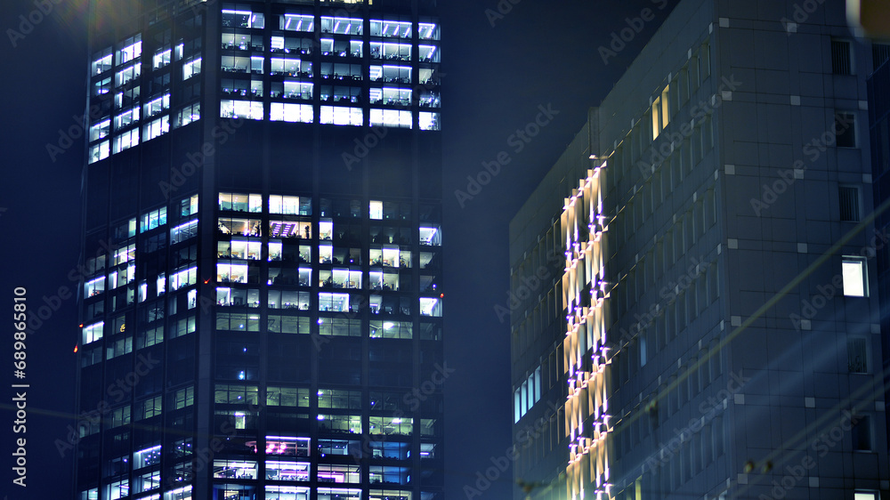 The glass facades of a modern skyscrapers at night. Modern glass office in city. Big glowing windows in modern offices buildings at night. In rows of windows light shines. The glass facades of a modern skyscrapers at night. Modern glass office in city. Big glowing windows in modern offices buildings at night. In rows of windows light shines.